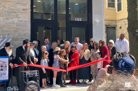 Group of Jews at a ribbon cutting ceremony where one person is cutting a large red ribbon
