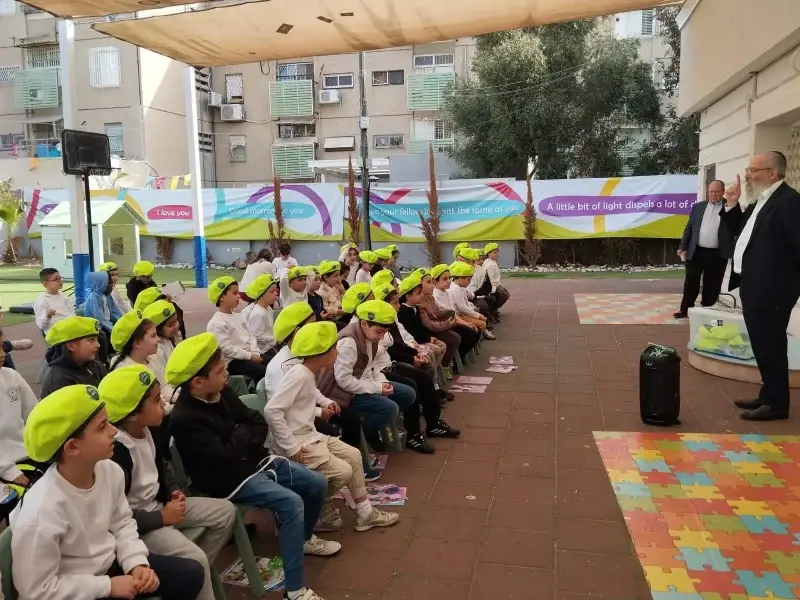 ילדים בכובעים צהובים יושבים בחצר המקורה ומקשיבים לדובר