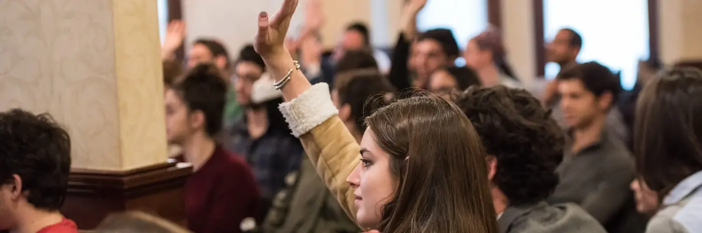 A woman with long brown hair raising her hand in a large conference room or audience setting.