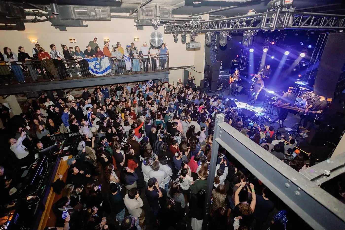 Crowd attending a live music concert in an indoor venue with stage lighting and performers playing guitars and drums.