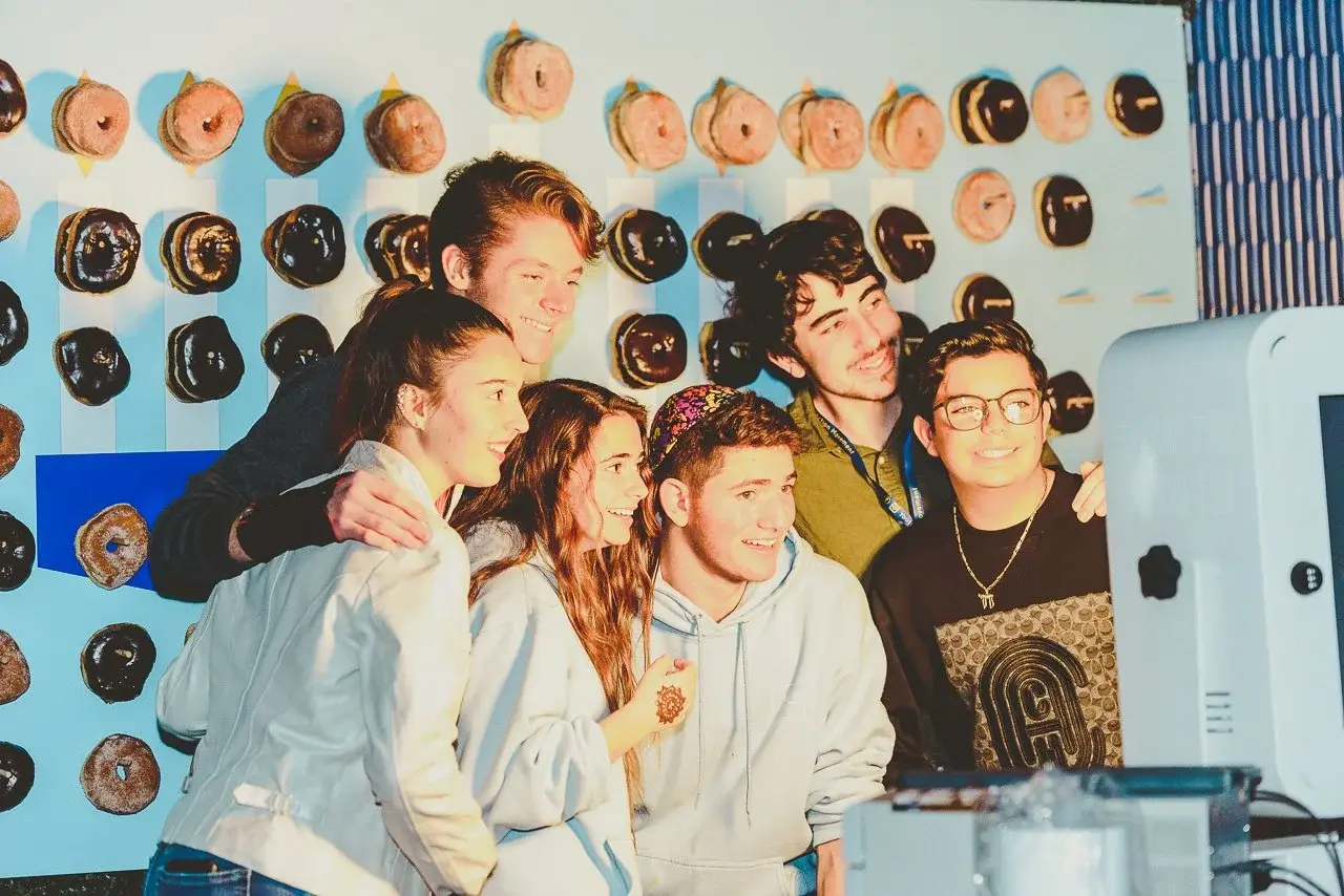 Group of six young friends taking a selfie in front of a wall decorated with donut-shaped objects.