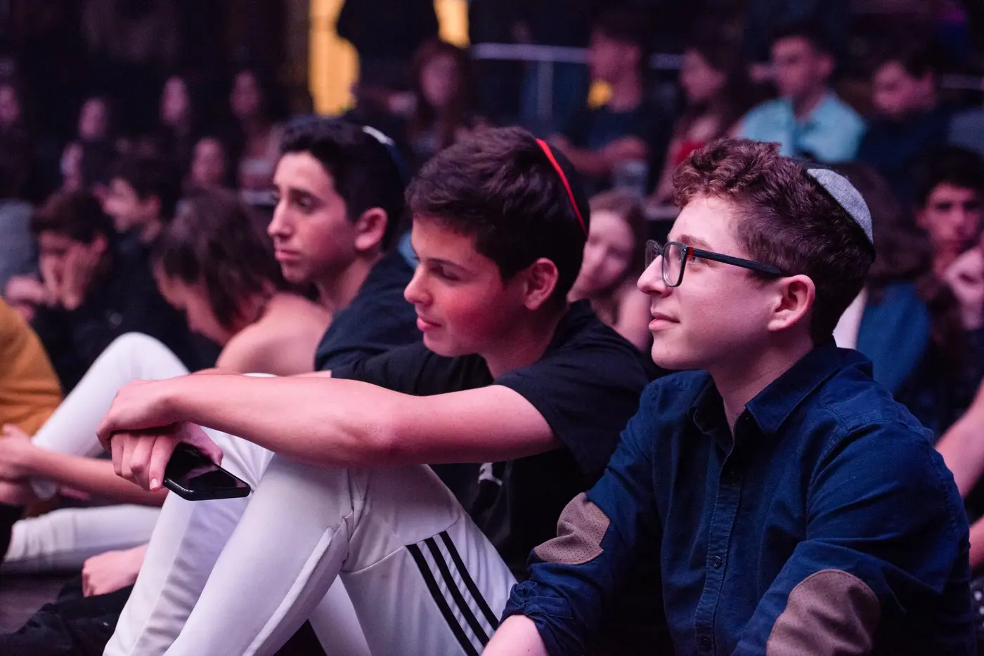 A group of young people sitting closely together in a dimly lit, indoor setting, listening attentively to a presentation or performance.
