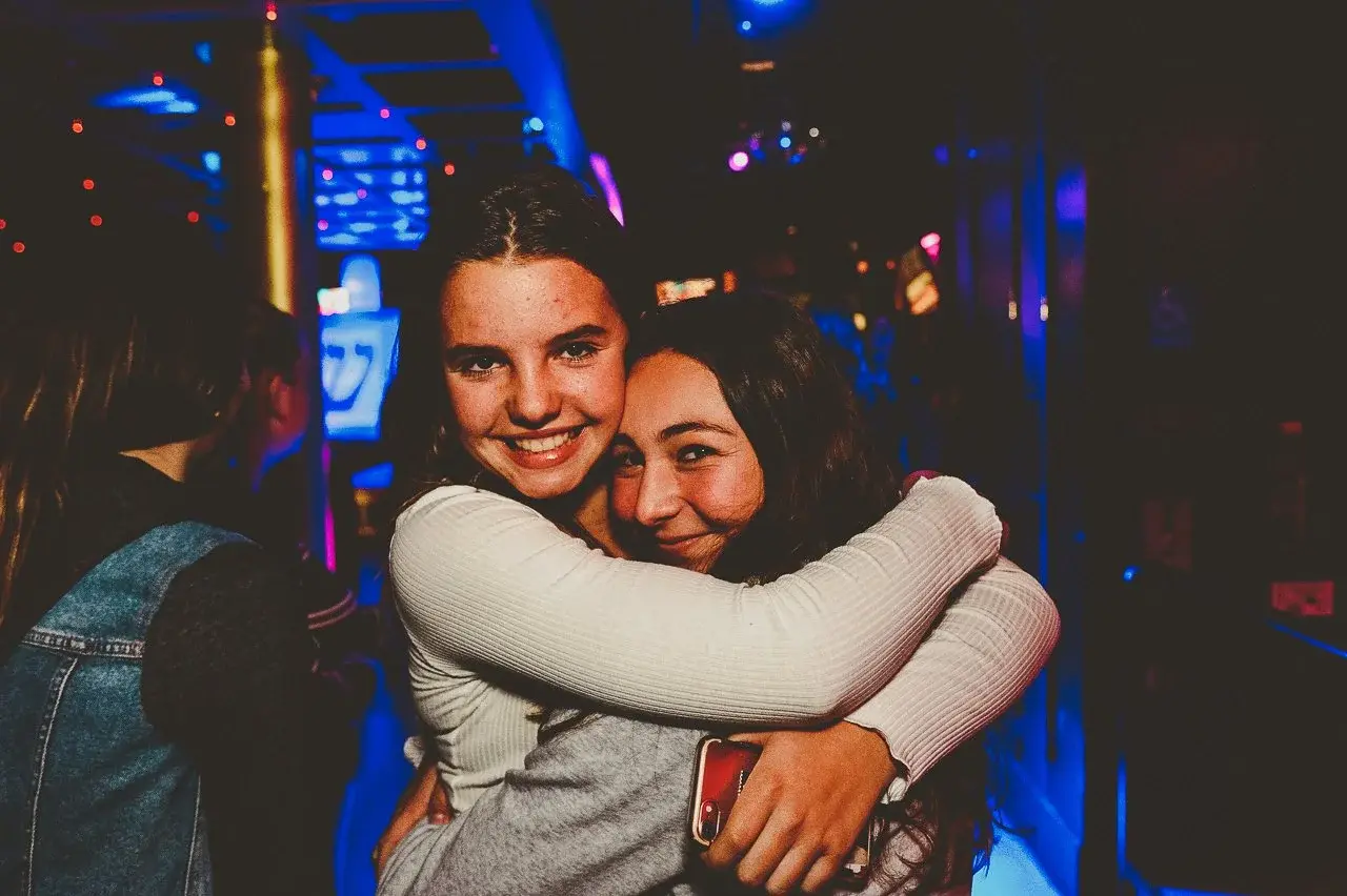 Two young women hugging and smiling in a nightclub with colorful neon lights in the background.