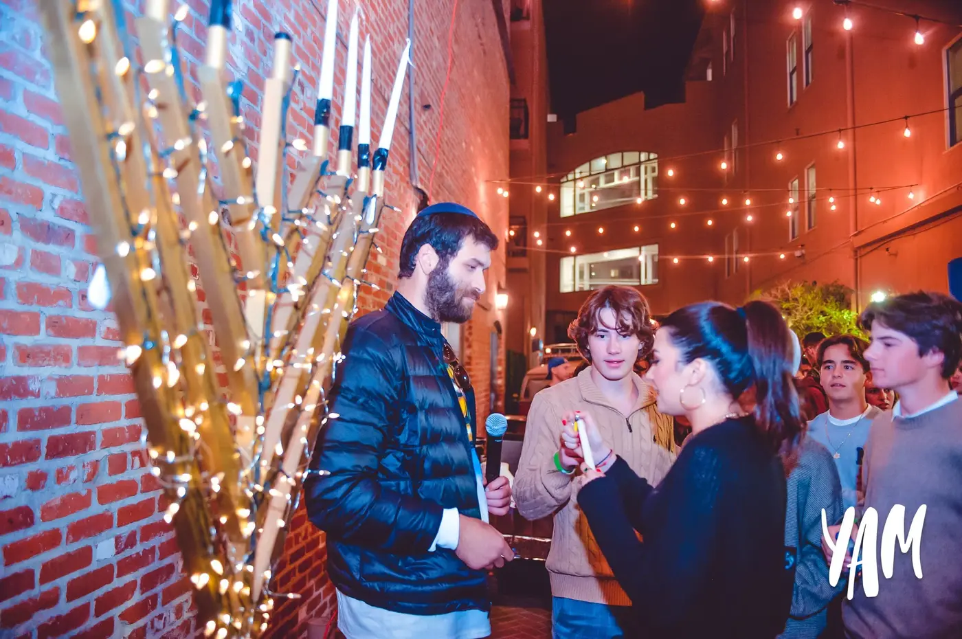 Group of young people talking at an outdoor evening event with string lights and brick walls.