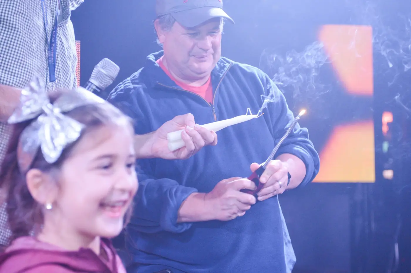 A man in a baseball cap holding a lit candle and a fork, with a young girl smiling in the foreground.
