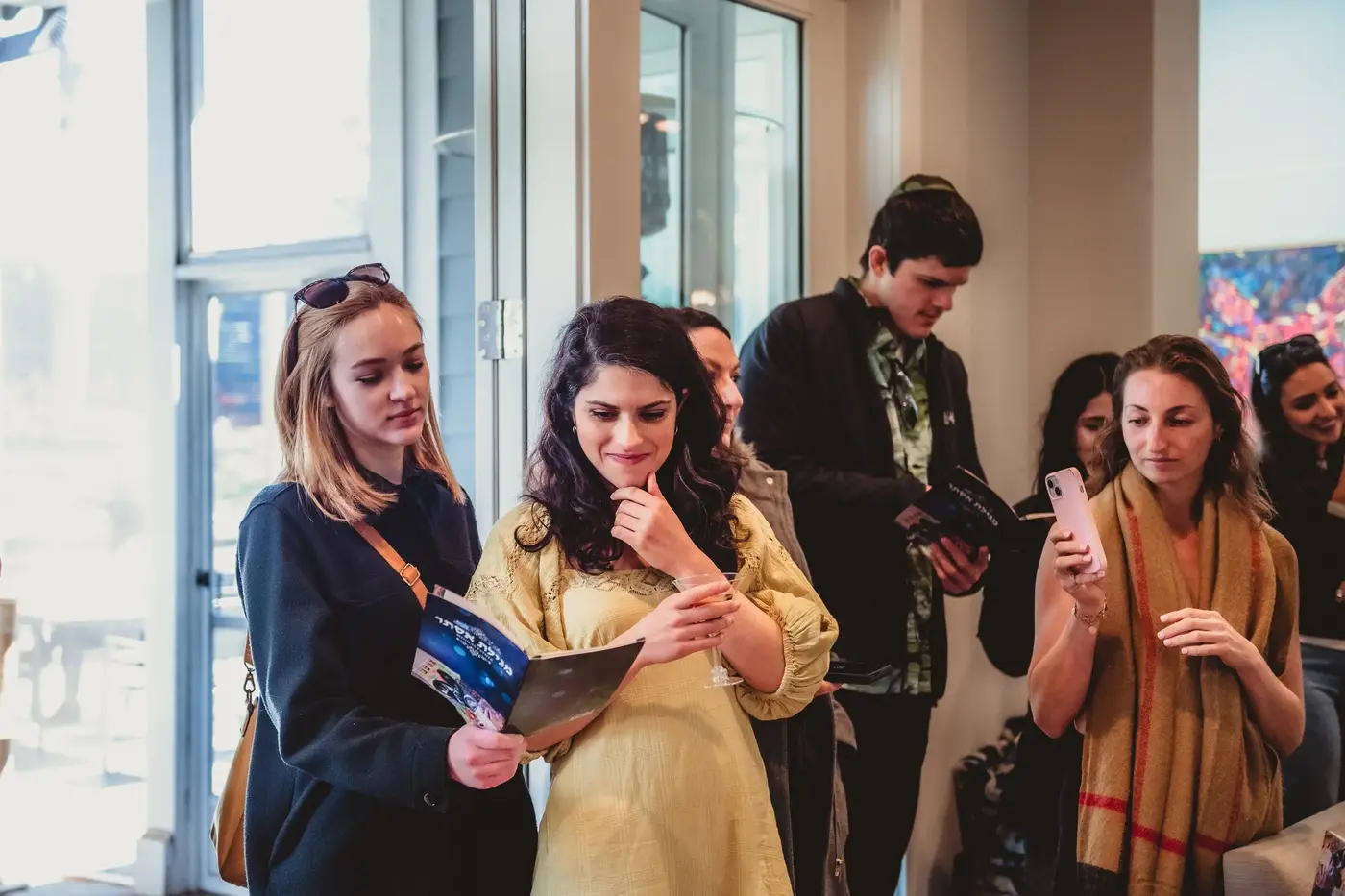 Group of people inside a room, some reading pamphlets, others using their phones, near a glass door with sunlight coming through.