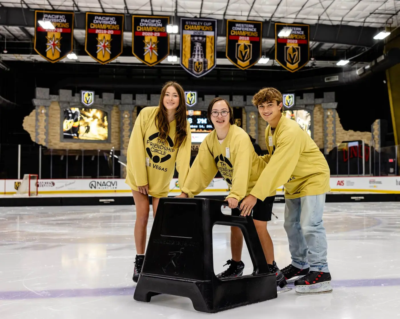 Three smiling young people in yellow "The Friendship Circle" shirts stand on an ice rink with a skating aid.