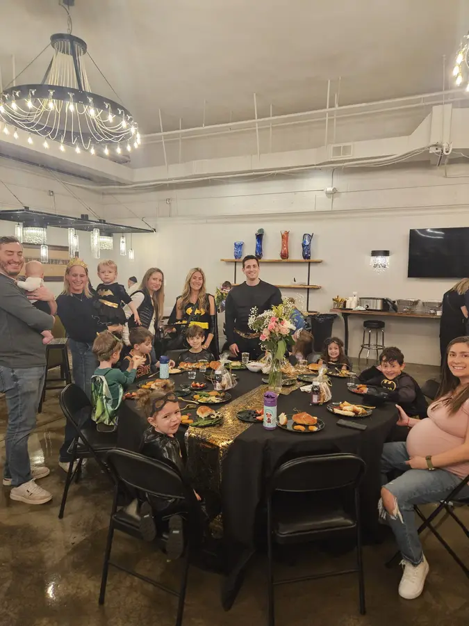 A group of children and adults gathered around a table with food at a party or celebration in an indoor venue with modern decor.