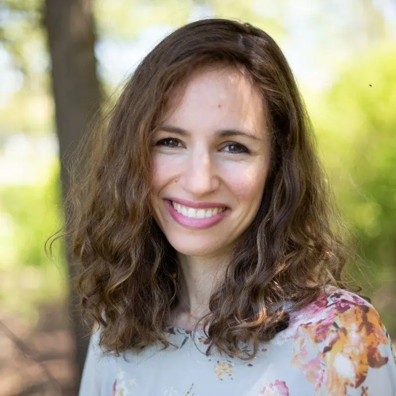 Smiling woman with curly brown hair, wearing a floral top, standing outdoors with blurred greenery in the background.