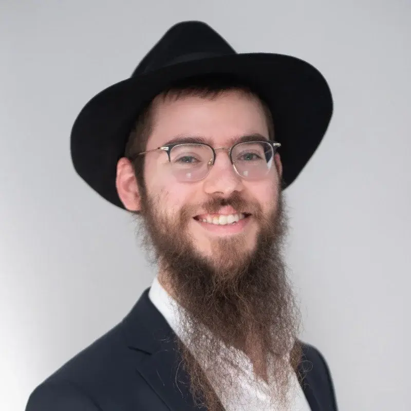 A man with a beard and glasses wearing a black hat and a dark blazer, smiling against a plain light-colored background.