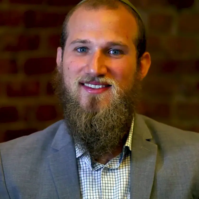 A man with blue eyes, a beard, and a mustache, wearing a gray suit and a checkered shirt, smiling in front of a brick wall.