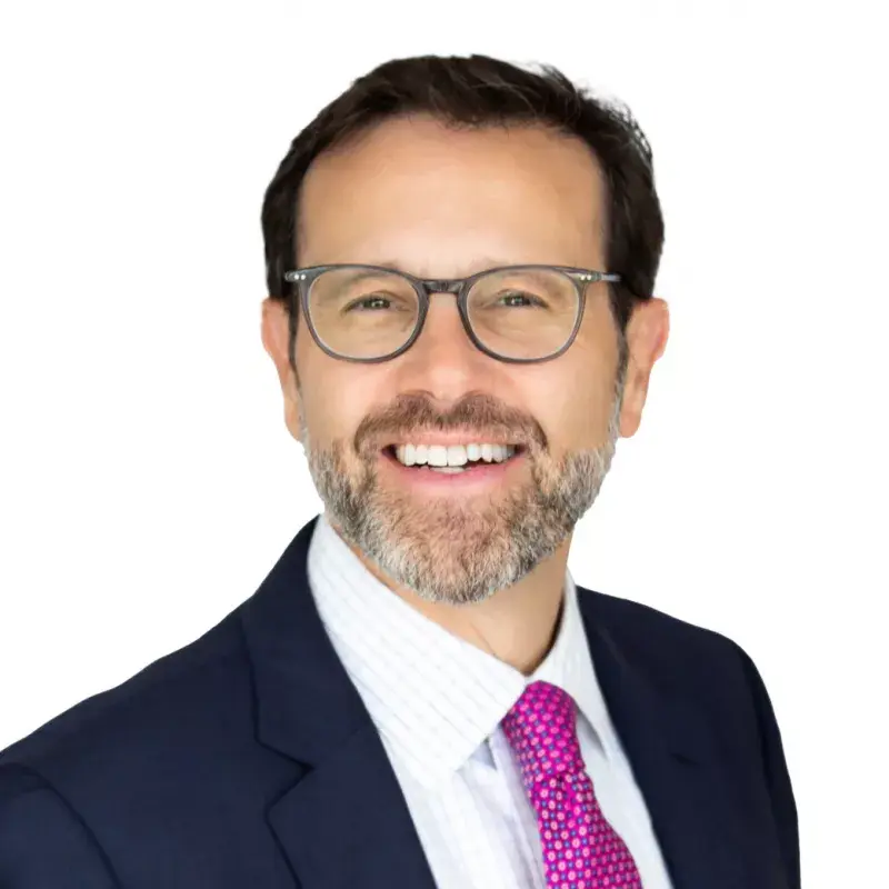 A smiling man with glasses, a beard, wearing a dark suit, white shirt, and pink tie.
