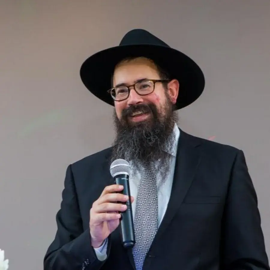 A man wearing glasses, a black hat, a dark suit, and a patterned tie, holding a microphone and smiling.