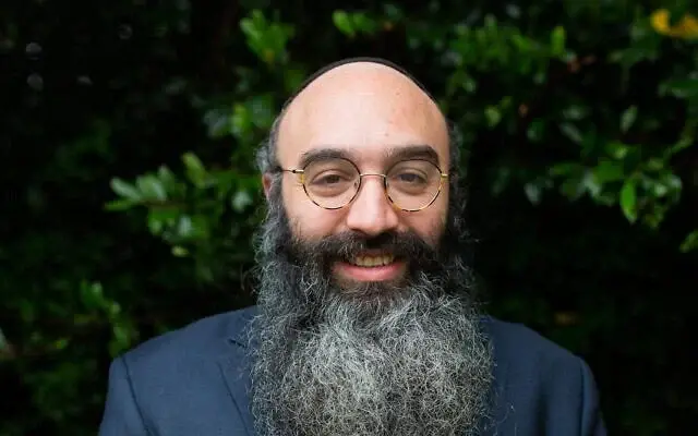 A man with glasses, a full beard, and a shaved head, smiling outdoors with green bushes in the background.