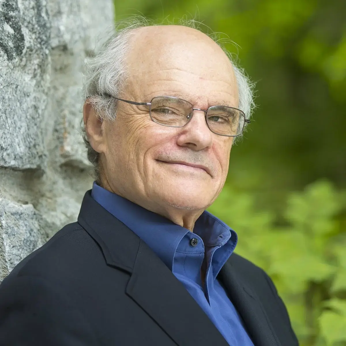 Portrait of an elderly man with glasses, smiling, in a black suit with a blue shirt, standing outdoors near a stone wall and greenery.