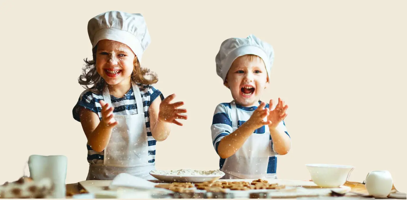 Two children wearing chef hats and aprons cooking and baking together, covered in flour with a kitchen table and baking ingredients.