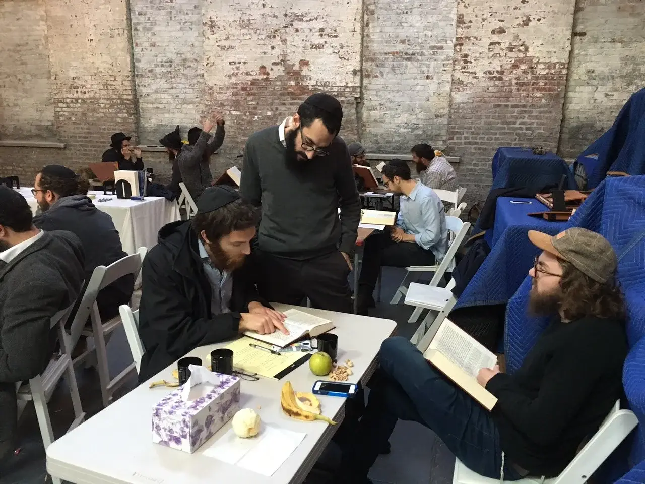 Several men studying or reading books in a room with exposed brick walls, some wearing hats, and a table with snacks and a tissue box.
