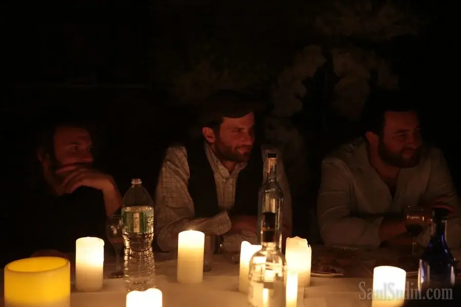 Three men sitting at a table with candles, water bottles, and glasses, in a dimly lit environment.