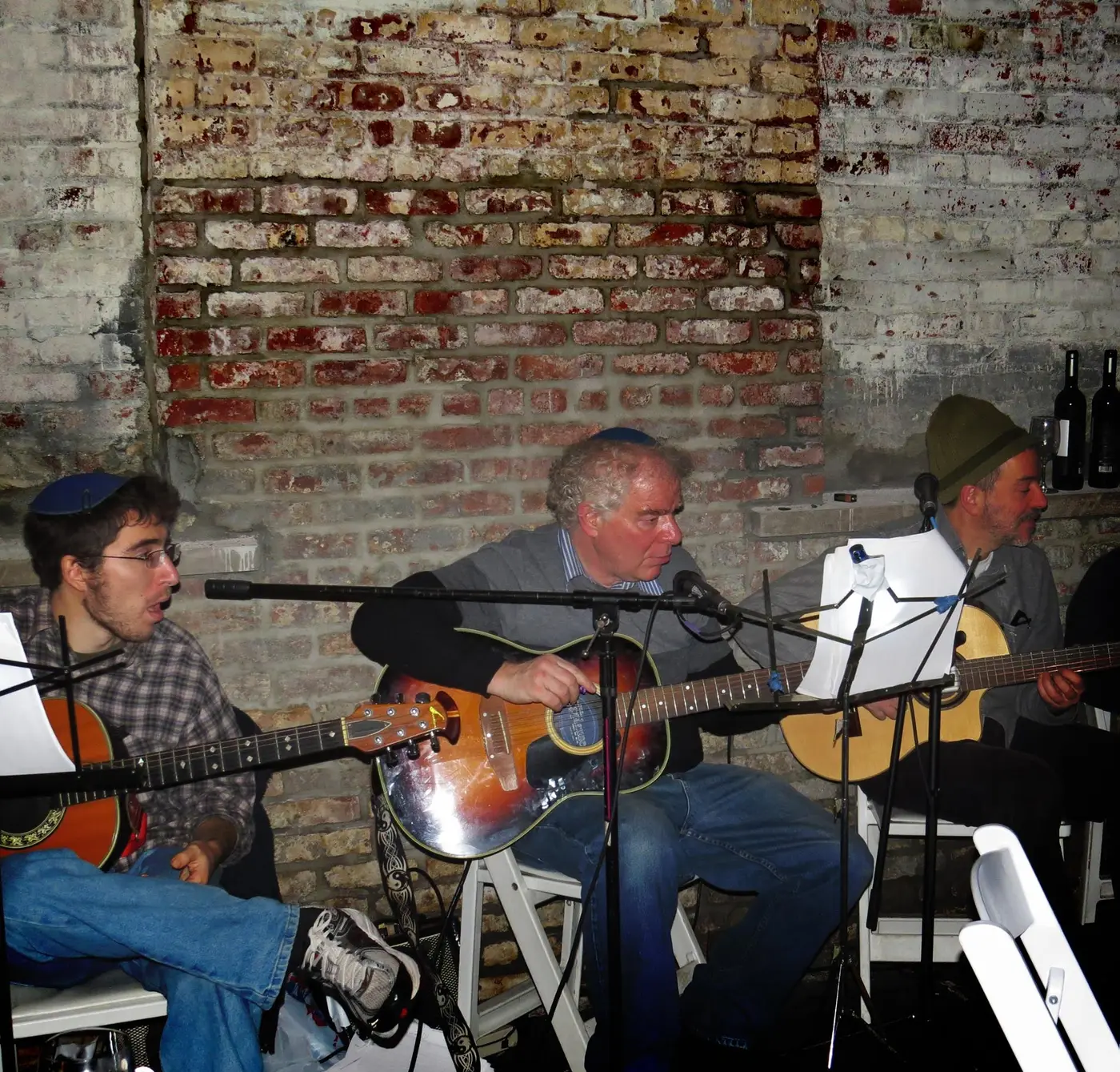Three men playing acoustic guitars in a room with exposed brick wall, music stands, and wine bottles.